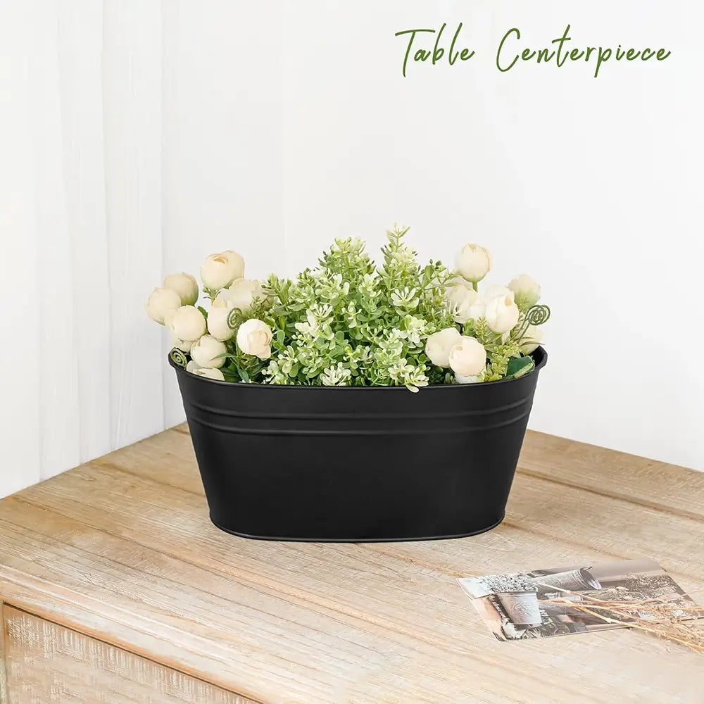 A black oval metal planter filled with faux white roses and green foliage, displayed on a light wood table against a white wall. The text 'Table Centerpiece' is written in green at the top right.