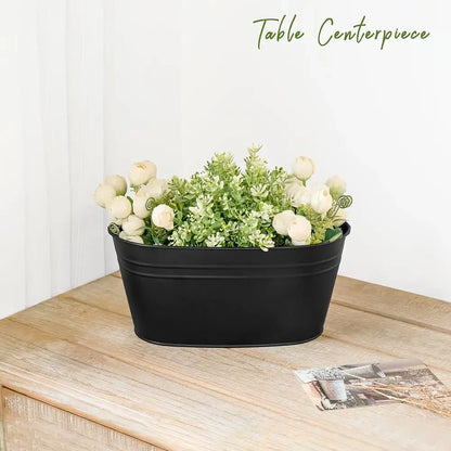 A black oval metal planter filled with faux white roses and green foliage, displayed on a light wood table against a white wall. The text 'Table Centerpiece' is written in green at the top right.