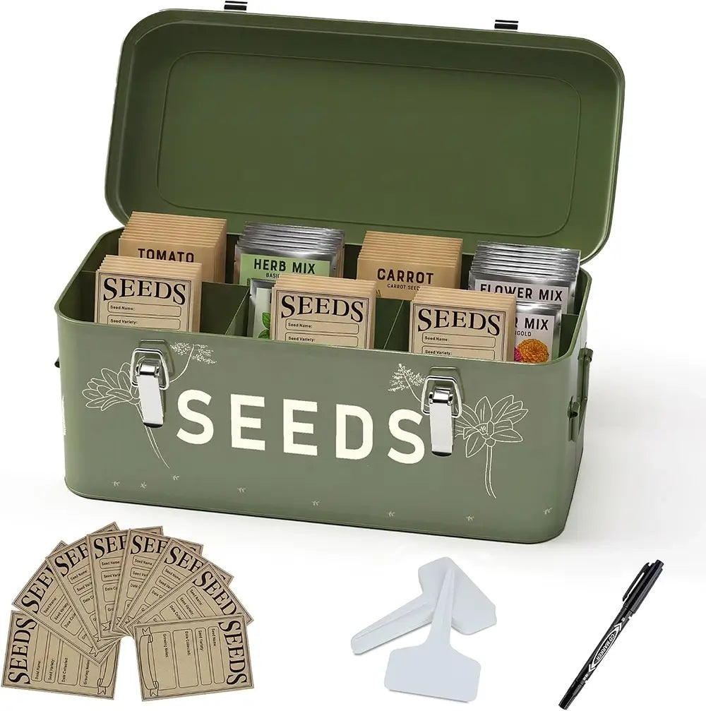An olive green metal seed storage tin with the word 'SEEDS' printed on the front, shown open with seed packets arranged inside. The tin is photographed on a clean white background with a few loose seed packets, two white spatulas, and a black pen placed in front.