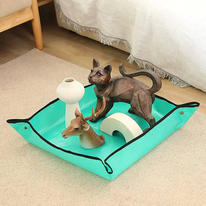 A turquoise pet play mat with black trim and handles, styled with decorative figurines including a cat, deer, and small white arches, set on a beige carpet in a home environment.