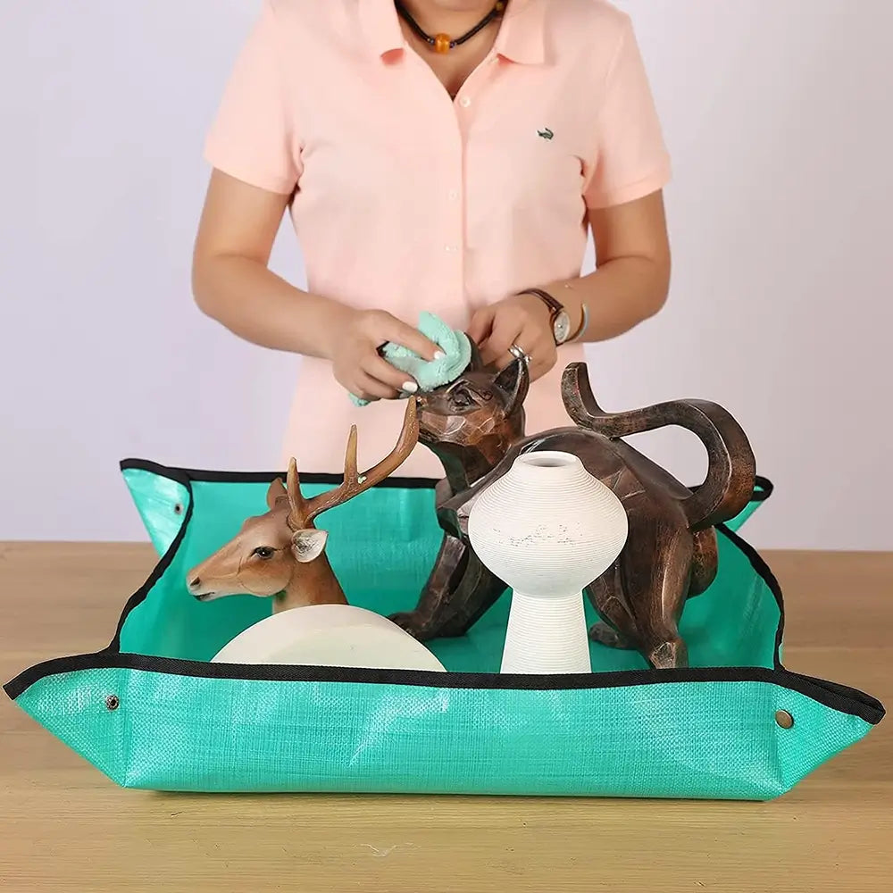 A person in a light pink polo shirt is shown from the chest down, holding a mint green cloth over a turquoise and black storage basket filled with decorative ceramic and wooden figurines, set on a light wood surface.