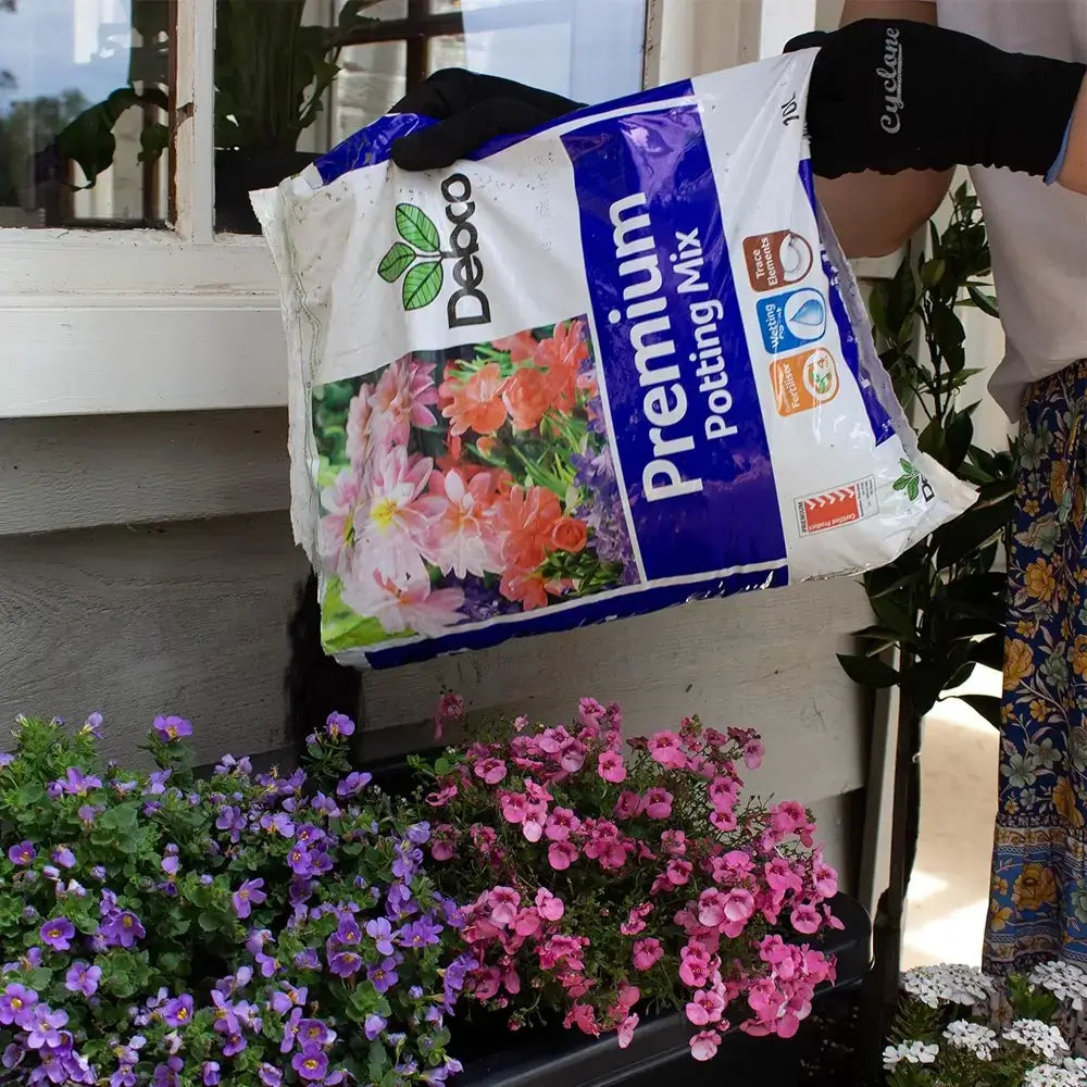 Person holding a bag of Debco Premium Potting Mix with flowers in the foreground